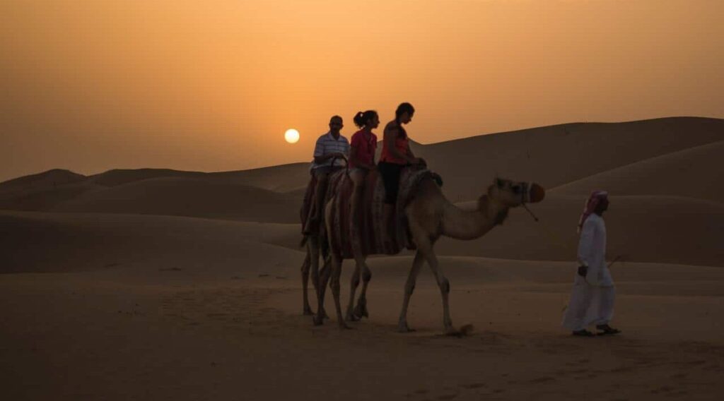 Three tourists riding two camels are guided through the desert by the camel keeper.